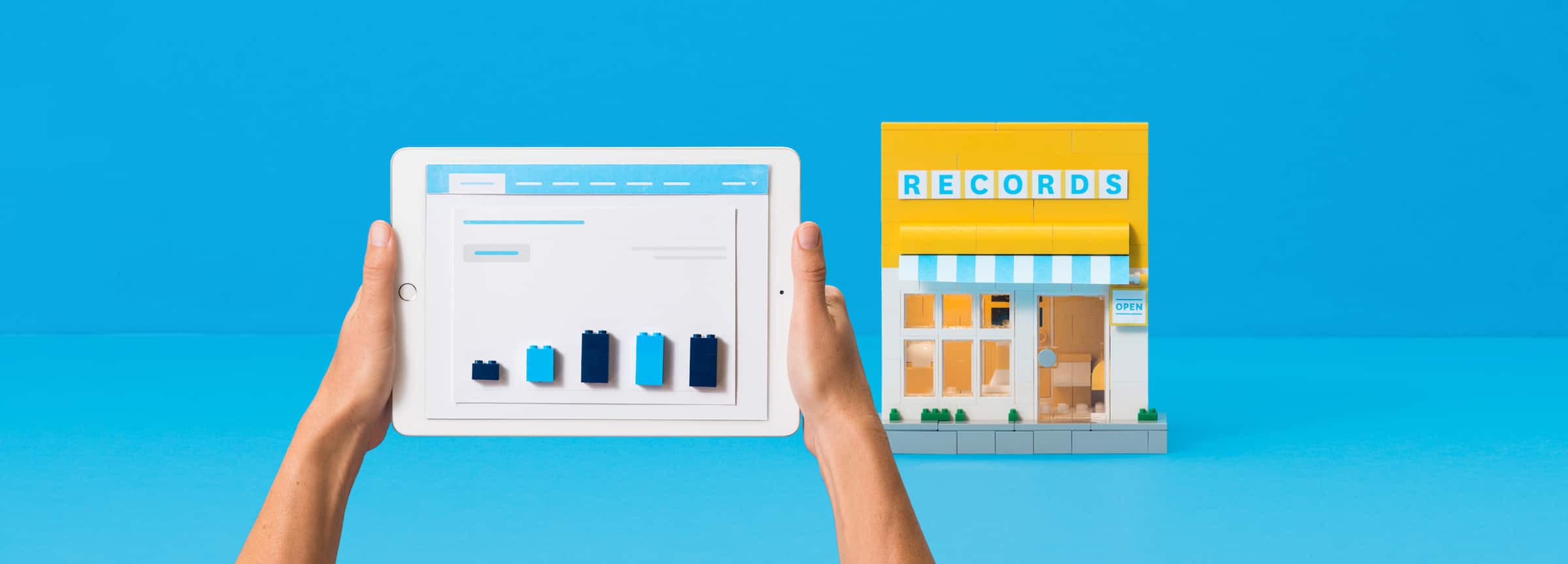 A person's hands holding a tablet, with a store made of lego in the background