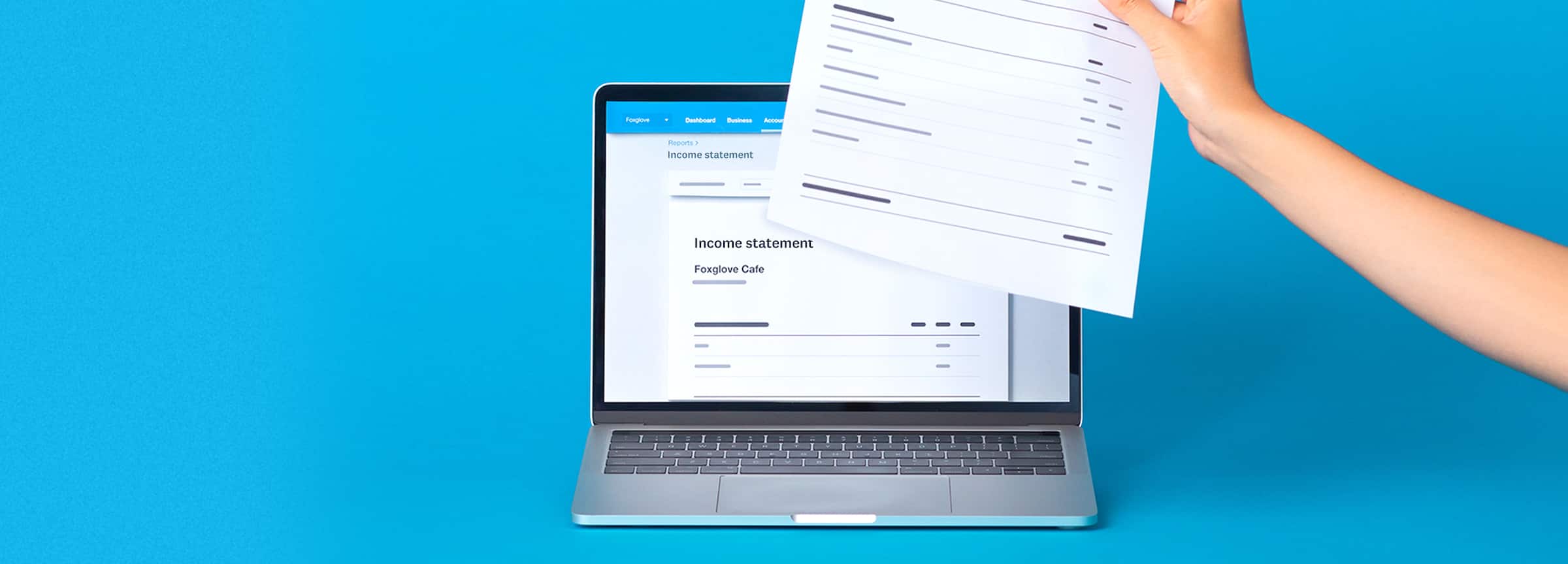 A hand holding an income statement in front of a laptop